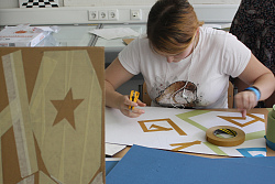 Zu sehen ist eine Schülerin mit Cuttermesser in der Hand. Sie sitz gebeugt über einem Karton, den sie mit Tape beklebt hat.