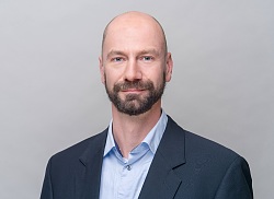 Portätfoto eines Mannes im mittleren Alter mit dunkelblauem Anzug und hellblauem Hemd. Er hat blaue Augen, kurz geschorenes Haar, trägt eine Vollbart und schaut in die Kamera.