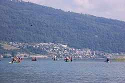 Das Bild zeigt mehrere Kanus auf einem großen See oder Fluss im Hintergrund am Ufer sind die Hochhäuser einer Stadt zu erkennen.