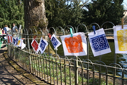 Das Bild zeigt zahlreiche bunte Werke, die mit Wäscheklammern an einer Schnur befestigt am Zaun um den Teich im Stadtpark hängen. 