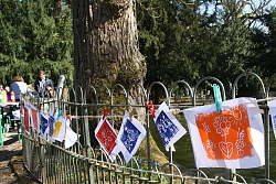 Das Bild zeigt zahlreiche bunte Werke, die mit Wäscheklammern an einer Schnur befestigt am Zaun um den Teich im Stadtpark hängen. 