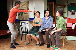 Das Bild zeigt eine Szene aus Achtung Deutsch!, die in einer Wohnung spielt. Ein Kühlschrank und ein Sofa stehen auf der Bühne. Ganz links steht ein Darsteller in grauer, langer Unterhose und rotem, langem Unterhemd mit Hauspantoffeln. Rechts von ihm sitzen auf Stühlen zu ihm gewandt drei weitere Darsteller, eine Frau im Dirndl und zwei Männer in Hose und Hemd beziehungsweise Polo-Shirt mit Hut. Der Mann in Unterhose zeigt auf den Mann ganz rechts mit Hut.   