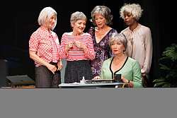 Das Bild zeigt eine der Sekretärinnen mit starrem Blick am Schreibtisch sitzen. Mit ihren Händen scheint sie ein Laptop auf oder zuzuklappen. Hinter ihr stehen vier Kolleginnen. Alle sind leicht skurril gekleidet. 