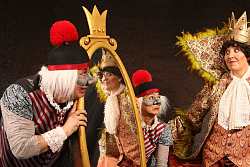 Das Bild zeigt den Kaiser, dargestellt von einer Frau, in einem prächtigen goldfarbenen Mantel mit einem an den Rändern federnbesetzten Stehkragen, der ihm bis über den Kopf reicht. Der Kaiser betrachtet sich in einem gekrönten Spiegel, der von zwei clown-artig angezogenen Dienern gehalten wird. 