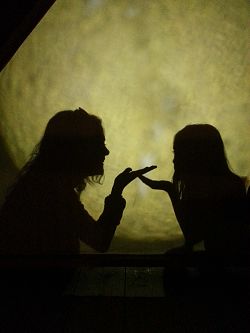 Das Bild zeigt die Schatten von einer Frau links und einem Mädchen rechts hinter einem Vorhang, die sich Küsschen zupusten.