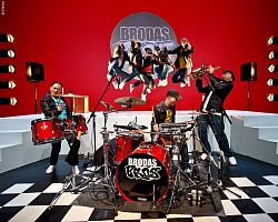 Das Bild zeigt drei Mitglieder der internationalen Hip Hop, Dance und Urban Culture Company an zwei Schlagzeugen und einem Saxophon. Auf der roten Wand im Hintergrund sind unter dem Schriftzug Brodas fünf in die Luft springende Tänzer zu sehen. 