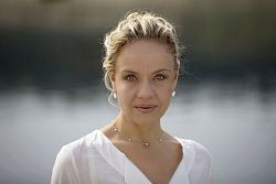 Das Bild zeigt ein Porträt von Anke Sieloff. Sie hat blondgefärbte, gelockte, hochgesteckte Haare und trägt eine weiße, kragenlose Bluse.