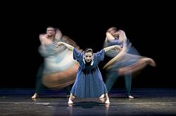 Das Bild zeigt eine Tanzszene. Im Vordergrund ist eine Frau im blauen Kleid mit breit geöffneten Beinen und Armen zu sehen.