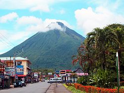 Das Bild zeigt eine breite Straße in einer Stadt am Rand sind Geschäfte zu erkennen, im Hintergrund erhebt sich ein riesiger, oben wolkenverhangener Berg.