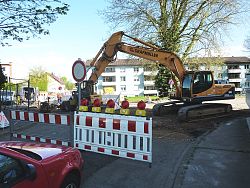 Das Foto zeigt die Baustelle zur Sanierung des Mischwassersammlers zur Kläranlage im Streckenabschnitt Kanadaring. Man sieht im Vordergrund ein rotes Auto, das direkt vor zwei rot-weißen Absperrzäunen steht, dahinter ein "Durchfahrt verboten" Verkehrszeichen und einen Bagger. Der Straßenbelag wurde abgetragen. Im Hintergrund sind Bauarbeiter zu sehen und die Häuser im Wohngebiet Kanadaring.