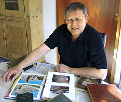 Das Bild zeigt Norbert Klein. Er sitzt an einem Tisch, auf dem etliche Bücher liegen sowie ein Album mit Fotos historischer Gebäude 