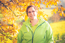 Portraitfoto von Katrin Kupferschmidt. Sie steht mit einem hellgrünem Polosghirt, über dem sie ein ebenfalls hellgrüne sportliche Jacke trägt, vor einem herbstlichen Hintergrund: ein Laubbaum mit gelb gefärbten Blättern und Wiesen. Kartin Kupferschmidt hat kurzes rötlich-braunes Haar und trägt eine schmale Brille.