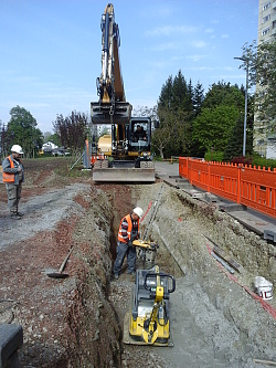 Ein Bagger hat einen Graben zum Verlegen des Kanals ausgehoben. Ein Bauarbeiter mit orangener Schutzweste und weißem Helm arbeitet in dem Graben. 