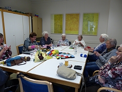 Auf dem Bild sieht man neun ältere Damen um einen großen Tisch sitzen, während sie ihre Handarbeiten verrichten. Es scheint eine gesellige Runde zu sein.