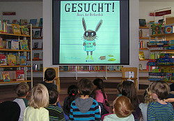 Das Bild zeigt ein Bilderbuchkino auf einer großen Leinwand in der Kinderabteilung der Mediathek. Ganz gebannt zusehend sitzen mit dem Rücken zu Kamera viele Kinder.
