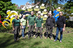 Lahrer Stadtgärtner stehen in Dole vor einem gestalteten Blumenbeet. Bunte Fahrradfahrer werden in dem Beet von Bienen verfolgt und zu schnellerem Fahren motiviert. 