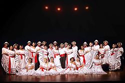 Gruppenbild des 31-köpfigen Chors aus Alajuela in ihren Kostümen. Das Bild ist auf einer Bühne gemacht. Der Hintergrund ist fast schwarz. Die Männer tragen schwarze Hosen, ein weißes Hemd, eine rote Schärpe um den Bauch und ein rotes Halstuch. Die Damen tragen lange weiße schulterfreie Kleider mit wenigen roten und schwarzen Streifen und Blumen in den Haaren. 
