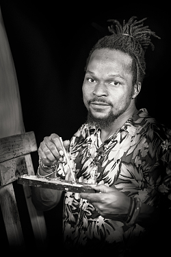 Zu sehen ist eines der Bilder aus der Fotoausstellung 'Das gehört zu mir'. Das Schwarz-Weiß Foto zeigt einen jungen dunkelhäutigen Mann mit einem Zopf aus Rastazöpfen, der mit Pinsel und Farbpalette vor einer Staffelei steht.