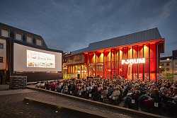 Vor dem hell in orange erleuchteten Forumkino sitzen Menschen in Stuhlreihen vor einer großen Leinwand auf dem Urteilsplatz. 