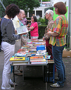 Beim Sommerabend der Mediathek Lahr bietet der Förderkreis der Mediathek einen Bücherflohmarkt. Das Bild zeigt einen Tisch voll Bücher im Freien, an dem Besucher schmökern und Mitglieder des Freundeskreises Rede und Antwort stehen. 