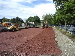 Auf dem Bild ist die Baustelle Parkplatz ortenauhalle zu sehen. Es wird gerade die Baugrundverbesserung aufgebracht. Man sieht einen Bagger und einige weitere große Bau- und Planierfahrzeuge bei der Arbeit.