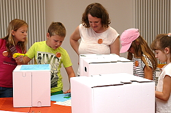 Beim Sommerferienprogramm des Stadtmuseum Tonofenfabrik kreieren Kinder Wellpappkartons. Sie bekleben die Kartons mit Papier und Motiven, ausgeschnitten aus alten Programmheften und Zeitungen. Museumpädagogin Silke Höllmüller unterstützt die Kinder. 