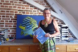 Martina Bauder, Geschäftsführerin von Taucher-Reisen, steht vor einem großen bunten Bild mit einem Fisch und hält ein Prospekt von atmosfair in den Händen. Sie hat kurzes graues Haar, einen sonnengebräunten Taint und trägt eine rote Kunststoffbrille. Sie hat eine bunt gemustete Hose mit Dschungelmotiven und ein schwarzes Top an, um den Hals trägt Sie eine Kette mit einer großen Muschel.