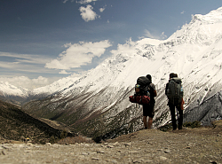 Die Rucksacktouristen Gwendolin Weisser und Patrick Allgeier stehen mit dem Rücken, bepackt mit dicken Rucksäcken, vor einem bergmassiv. Oben sind die Spitzen mit Schnee bedeckt. 