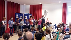 Einige Kinder stehen in einer Reihe auf der Bühne und halten jeweils ein Blatt mit einem Buchstaben vor den Körper.