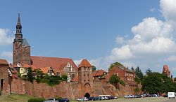 Zu sehen ist eine Stadtansicht von Tangermünde in der Altmark mit einer alten Stadtmauer, einer Kirche und roten Ziegelgebäuden.