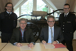 Die Feuerwehr Rust beteiligt sich am Schlauchpool der Stadt Lahr. 
An einem Tisch sitzen Oberbürgermeister Dr. Wolfgang G. Müller (links) und Rusts Bürgermeister und haben die Verträge vor sich. Sie lächeln beide in die Kamera. Umrahmt werden sie von ihren Feuerwehrkommandanten, die jeweils schräg in Uniform dahinter stehen. Im Hintergrund sieht man eine historische Feuerwehrpumpe.