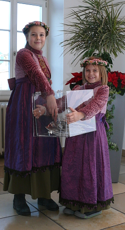 Zwei Mädchen in violettfarbenen Kleidern mit Blumenkränzen auf dem Kopf. Ein Mädchen hält einen transparenten Behälter mit Postkarten, das andere Mädchen zieht Postkarten daraus.
