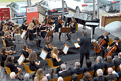 Die Sinfonietta tritt bei der Veranstaltung Musik.Genuss auf.
Das Bild zeigt die Musiker, schwarz gekleidet, von der Perspektive von oben. Das Bild zeigt die Streicher im Halbrund. Im Hintergrund ist eine Glaswand, die einen Blick auf abgestellte Audis zulässt. Zudem ist die erste Reihe der Zuhörer von hinten zu sehen. 