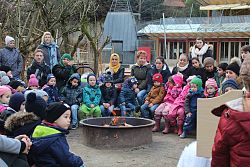 Kinder und Eltern sitzen um ein Lagerfeuer im Winter. Alle sind mit dicken Jacken, Schals und Mützen dick eingemummelt. 