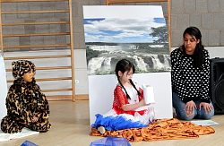 Ein Kind sitzt vor einem Bild mit einem Wasserfall und öffnet eine Flaschenpost. Links von ihm sitzt ein weiteres Kind als Tiger verkleidet. Daneben kniet rechts eine Erzieherin. 