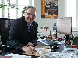 Erster Bürgermeister Guido Schöneboom am Tag nach der Wiederwahl an seinem Schreibtisch. Er trägt ein blaues Hemd, eine gestreifte Krawatte und einen dunklen Anzug mit einem Anstecker der Landesgartenschau Lahr 2018 und lacht in die Kamera.