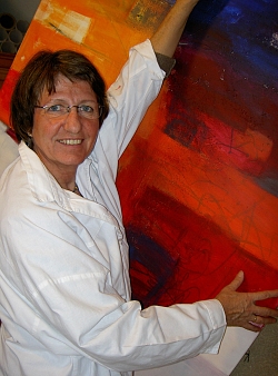 Frau mit braunen Haaren und weißem Kittel hält ein rot-orangefarbenes Gemälde in die Höhe und lächelt in die Kamera