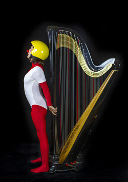 Eine Clowin lehnt sich an eine Harfe und streckt den Kopf nach oben. Sie trägt eine rote Clownnase und einen gelben Motorradhelm. Sie ist mit roten Leggins und und einem roten Shirt gekleidet, über die sie einen kurzen weißen Turnanzug trägt. 