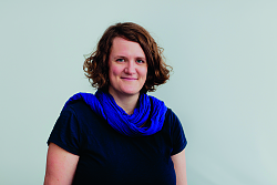 Porträt der Museumpädagogin Silke Höllmüller mit schwarzem T-Shirt und blauem Schal. Sie lächelt in die Kamera mit geschlossenem Mund und trägt einen gelockten Bob mit dunkelblondem Haar. 