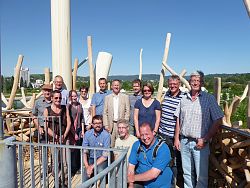 Zu sehen ist ein Gruppenfoto der Mitglieder der AG Saatkrähe bei einem Besuch der Landesgartenschau in Lahr. Die Gruppe von 14 Personen, gemischt Männer und Frauen, stehen im sogenannten "Krähennest" auf der LGS in Lahr. Es handelt sich um einen Turm, getragen von Holzpfeilern und einer Wendeltreppe aus Metall, dessen Aussichtsplattform gestaltet ist wie ein Vogelnest. Es wurden rundherum Hölzer angebracht, die den Eindruck eines übergroßen Nestes vermitteln. Im Hintergrund ein schönes Blick bei herrlichem Wetter über Lahr zum Schutterlindenberg.
