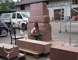 Die Künstlerin hat vier Sandsteine, nach oben kleiner werdend, übereinander gestapelt und bearbeitet knieend den unteren Sandstein mit einem Meisel. 