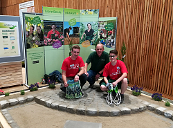 Die erfolgreichen Azubis der Stadt Lahr sitzen in der Hocke auf einem halbrund gepflasterten Platz in der Mitte ihr Chef Herbert Schneider. Die Azubis haben ein rotes T-Shirt an, Schneider ein grünes. Alle Drei lächeln in die Kamera. 