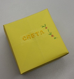 Das Bild zeigt eine gelbe Schachtel mit aufgeklebten Buchstaben, die den Namen Greta ergeben. Außerdem ist rechts auf die Schachtel ein kleiner Blumenzweig mit roten Blüten gemalt. 