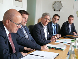 Blick ins Plenum der Pressekonferenz. Die Herren sitzen an einem langen Tisch nebeneinander, hören sich zu und sprechen. Alle sind mit Anzug gekleidet. 