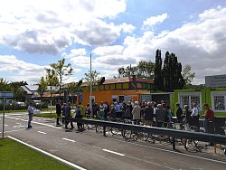Blick auf den Platz der Jugendverkehrsschule. Man sieht eine Straße mit Markierungen und dem Radsymbol, dazu verschiedene Verkehrszeichen und eine Leitplanke. Hinter dieser sind mehrere grün-gelbe Fahrräder geparkt und es stehen viele Menschen dort, die ihre Aufmerksamkeit auf den Erster Bürgermeister Guido Schöneboom gerichtet haben, der gerade spricht.