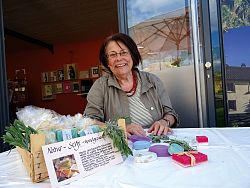 Das Bild zeigt Monika Fischer. Sie sitzt strahlend an einem Tisch, vor ihr liegen verschiedene Seifenprodukte und Seifenförmchen.