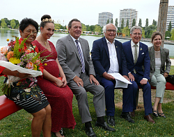 Auf einer geschwungenen roten Bank im Seepark der Landesgartenschau sitzen von links Frau Herrenknecht, Chrysanthemenkönigin Lea Johanna I., Altbundeskanzler Gerhard Schröder, Martin Herrenknecht, Oberbürgermeister Dr. Wolfgang G. Müller und seine Ehefrau Elke Oberg. 