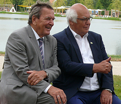 Auf einer geschwungenen roten Bank sitzen nebeneinander Gerhard Schröder und Martin Herrenknecht. Herrenknecht zeigt den erhobenen Daume und blicken beide nach rechts. 