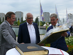 OB Dr. Müller (rechts) überreicht ein Geschenk an Herrn Herrenknecht. Gerhard Schröder steht links daneben. Im Hintergrund sind der Seepark und die Hochhäuser zu sehen. 