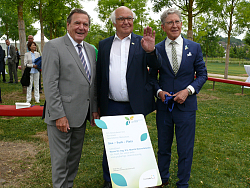 Tafel, die an die Widmung von Herrn Herrenknecht erinnert. Steht im Seepark und ist mit einem blauen Band umwickelt. 
Gerhard Schröder, Martin Herrenknecht winkend und Oberbürgermeister Dr. Müller stehen lachend dahinter. 